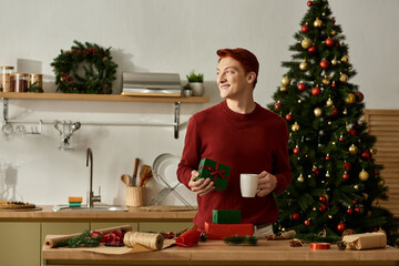 A young man holds a cup and a gift in a decorated kitchen, embracing holiday cheer and warmth.