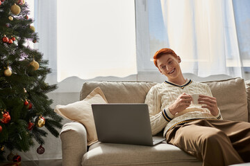 A young man smiles while seated on a couch, sipping a warm drink, surrounded by festive decor.
