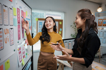 Two enthusiastic female web designers collaborating on a user interface design project, using a whiteboard and sticky notes in a modern office