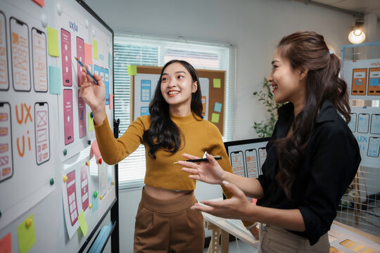 Two enthusiastic female web designers collaborating on a user interface design project, using a whiteboard and sticky notes in a modern office - Powered by Adobe