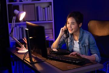 Young beautiful Asian creative woman calling on phone desk to customer or coworker explaining creator project's high profit while searching market analysis on pc at neon office at night. Stratagem.