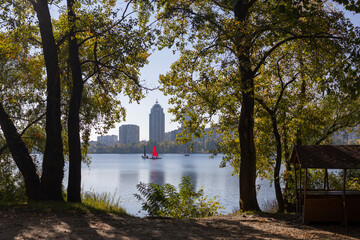 A serene view of a lake framed by trees, with red sailboats and a cityscape in the background under a clear sky