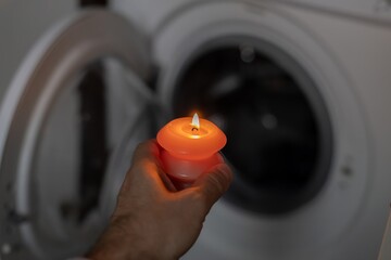 Burning candle in hand near washing machine in the dark. Power outage due to war in Ukraine