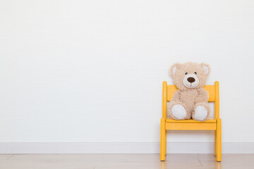Smiling brown soft teddy bear sitting on yellow small baby chair on wooden floor at light gray wall...