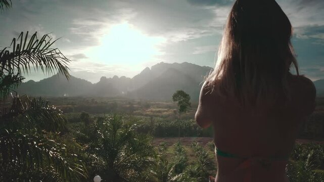 Woman enjoying the view from a wooden terrace in thailand, surrounded by lush greenery and mountains, under warm sunlight