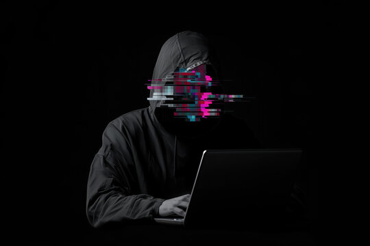 Dark person in a hood with a glitch instead of a face sitting at a laptop, cyber fraud and scam concept