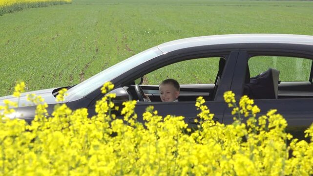 Happy little boy sits on driver seat in the car stopped in the yellow rape flowers field, listening to music, jumping and having fun