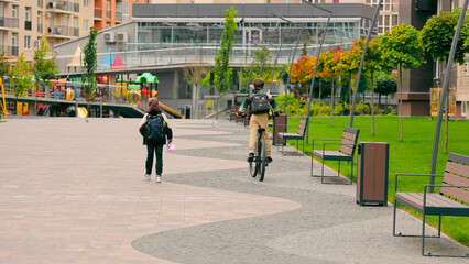 Two schoolchildren walking and biking through a modern park. Two school-aged children are seen from...