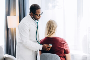 Fototapeta premium African American male doctor in glasses listening with stethoscope to female patient heartbeat