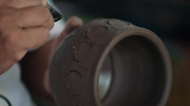 Close up cutter stained with clay in hands of careful professional adding minimalist design at pot before baking. Unknown artist sculpting ceramic utensil for selling unique piece for interior.