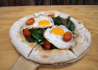 Closeup view of a pizza with mozzarella cheese, spinach, cherry tomatoes and fried eggs, on the wooden table.	
