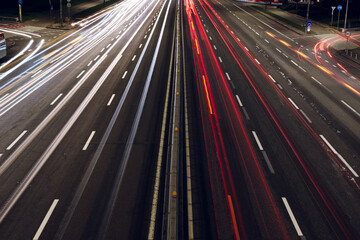 High-angle nighttime long-exposure shot of a highway, showing only the road and blurred car lights. Red and white light trails streak across the asphalt, capturing the dynamic energy of urban life.