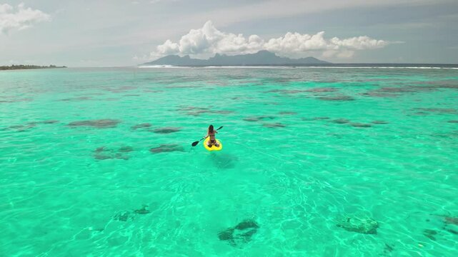 Woman is enjoying a sunny day on a yellow paddle board, surrounded by the turquoise water of a tropical lagoon with mountains in the background