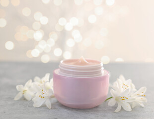 Cream in jar and beautiful jasmine flowers on grey table against light background with blurred lights, closeup. Space for text