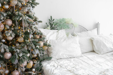 Cozy bedroom corner decorated for holidays with festive Christmas tree with golden ornaments, garland with lights and bed with white bedding and soft pillows, creating cozy holiday atmosphere.