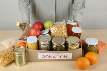 Food donation. Woman with box of different products at wooden table indoors, closeup