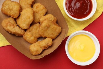 Delicious chicken nuggets in carton box and sauces on red background, flat lay