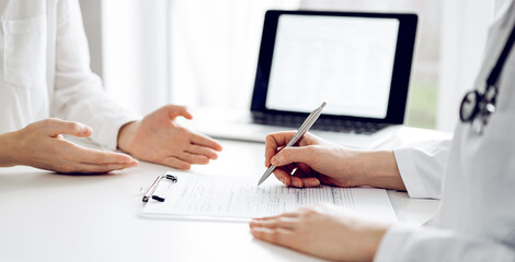 Doctor and patient sitting and discussing something near each other at the white desk in clinic. Female physician is listening filling up a records form. Medicine concept