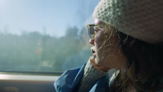 Young woman with knit hat travels by train looking outside and talking on phone. Female using smartphone enjoying conversation while she commutes to work or student going to university as view changes
