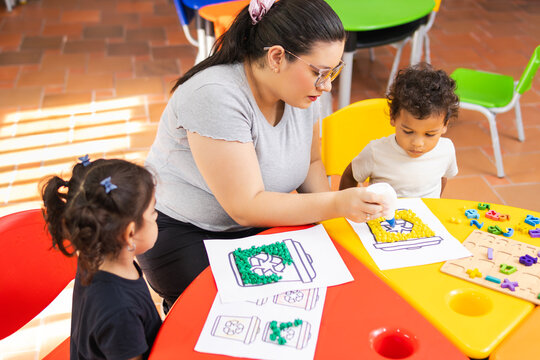 A kindergarten teacher assists her students with a recycling project, using waste bins for sorting materials
