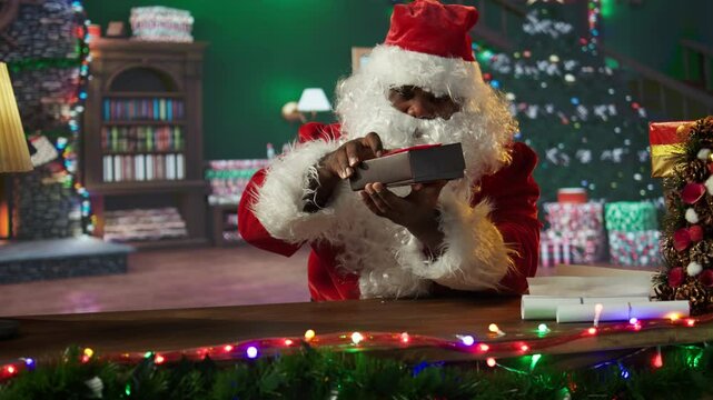 Black Santa wraps a festive gift box surrounded by Christmas lights and ornaments in his workshop. Father Christmas prepares for the holiday season, packing presents to bring joy. Camera B.