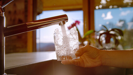 LENS FLARE, CLOSE UP: Hand holds and fills a glass with drinking tap water in modern kitchen. Crystal clear and fresh water splashes and glistens while the glass is filled in bright morning sunlight.
