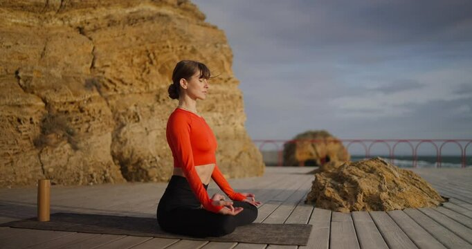 Side view of a happy brunette girl yoga master in an orange sweater sits in the lotus position and meditates on the beach covered with boards in front of the sea in the morning in summer
