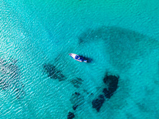 A single boat floats on clear, turquoise waters near Sardinia, Italy. Dark patches of underwater rocks or seaweed create a striking contrast.