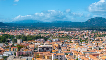 Naklejka premium Aerial view of Olbia, Sardinia, featuring terracotta roofed buildings, lush greenery, and mountains under a bright blue sky, showcasing urban and natural elements.