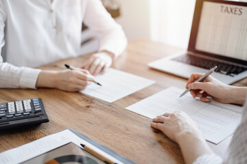 Woman accountant using a calculator and laptop computer while counting taxes with colleague at wooden desk in office. Teamwork in business audit and finance