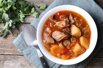 Delicious stew with vegetables in bowl, parsley and spoon on wooden table, top view