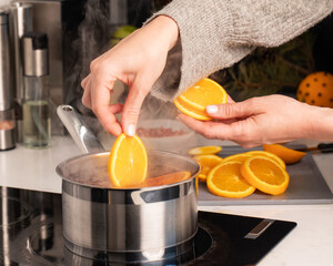 A woman adds a slice of orange to the boiling mulled wine in a saucepan in the kitchen.