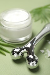 Metal face roller and cosmetic product on green background, closeup