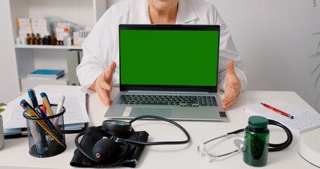 Green screen laptop mockup doctor explaining in white gown while sitting in health clinic. Doctorate having online conference.