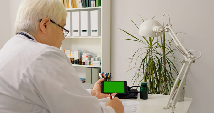 Over the sholder nurse writing in her notebook while watching green screen phone in office. Gadgets being used in medical service.