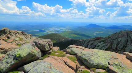 Panoramic view from a mountain peak, showing a valley with a lake and green hills in the distance. The foreground is rocky and covered with green moss.
