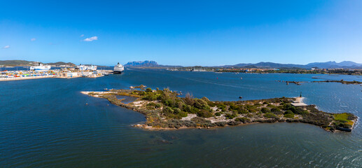 Obraz premium Aerial view of Olbia port in Sardinia, featuring cruise ships, a lush island, and the Mediterranean Sea, with rugged mountains in the background.