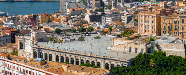 Fototapeta premium Aerial view of Cagliari, Sardinia, highlighting Bastione di Saint Remy with its terrace and arches. Colorful buildings and a bustling harbor are visible.