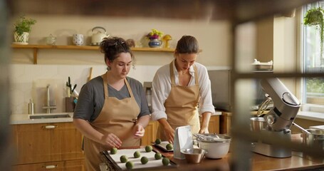 Confident girl with curly hair in a beige apron and her colleague Baker girl in a white shirt lay out green rolls of dough on a baking sheet while preparing dessert in a cafe