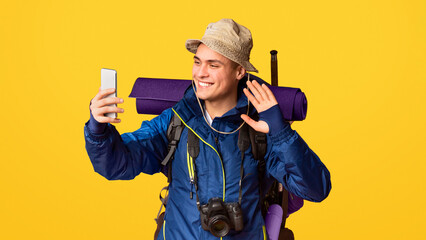 Smiling hiker in jacket with big backpack taking selfie on smartphone over orange studio background