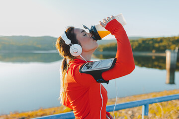 A woman in athletic attire is quenching her thirst with a bottle of orange juice, surrounded by the serene backdrop of a lake, wearing headphones and armband.