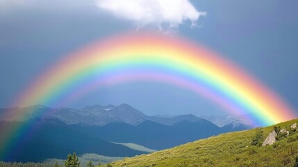 A vibrant rainbow arches over a range of mountains, with a clear blue sky and fluffy white clouds in the background.