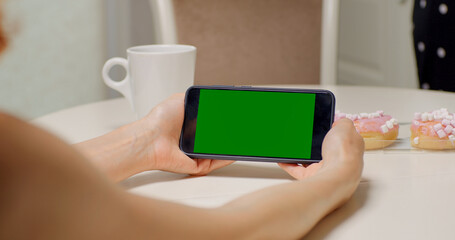 Close up shot of womans hands holding a green screen chroma key horizontal phone. Woman in the kitchen looking for recipe on green screen smartphone.