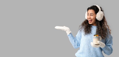 Happy young African-American woman holding cup of hot mulled wine and showing something on grey...