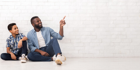 Great Offer. Cheerful Afro Man Sitting With His Son On Floor Near Brick Wall And Pointing At Copy...