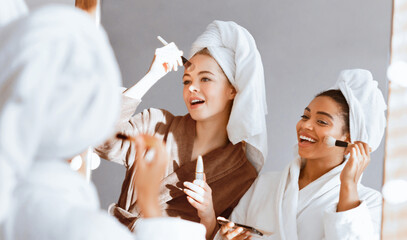 Cheerful multiethnic girls in bathrobes standing in front of big mirror, applying make up...