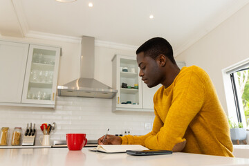 African american man in kitchen writing in notebook with smartphone and red mug nearby, at home
