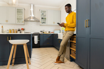 African american man in kitchen using smartphone, wearing yellow sweater, at home