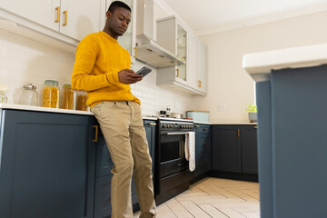 African american man in kitchen using smartphone, wearing yellow sweater, at home