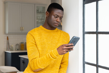 African american man in yellow sweater using smartphone by window at home, looking focused